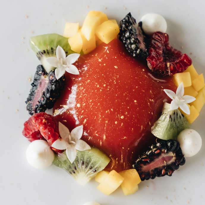 A close-up image of an artistic and colorful dessert plate, beautifully arranged with slices of mango, kiwi, blackberries, raspberries, and decorative white flowers, centered around a bright red, jelly-like dessert.