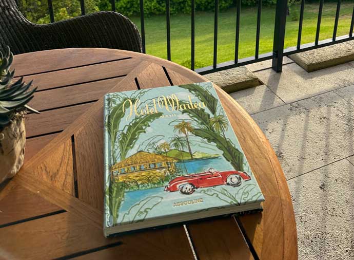 A photograph of a wooden outdoor table featuring the Hotel Wailea coffee table book. The table is on one of the suites' lanais overlooking a lush green lawn.