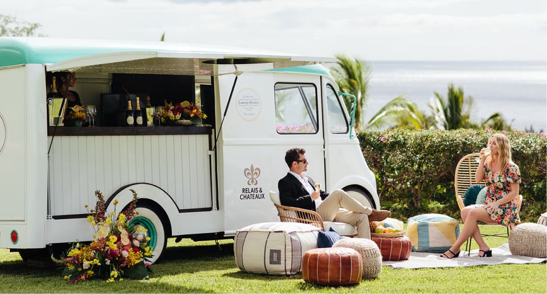 Two hotel guests enjoying the Relais & Chateaux Bubble Bus during an event in the bright Maui sunshine on top of cushions.