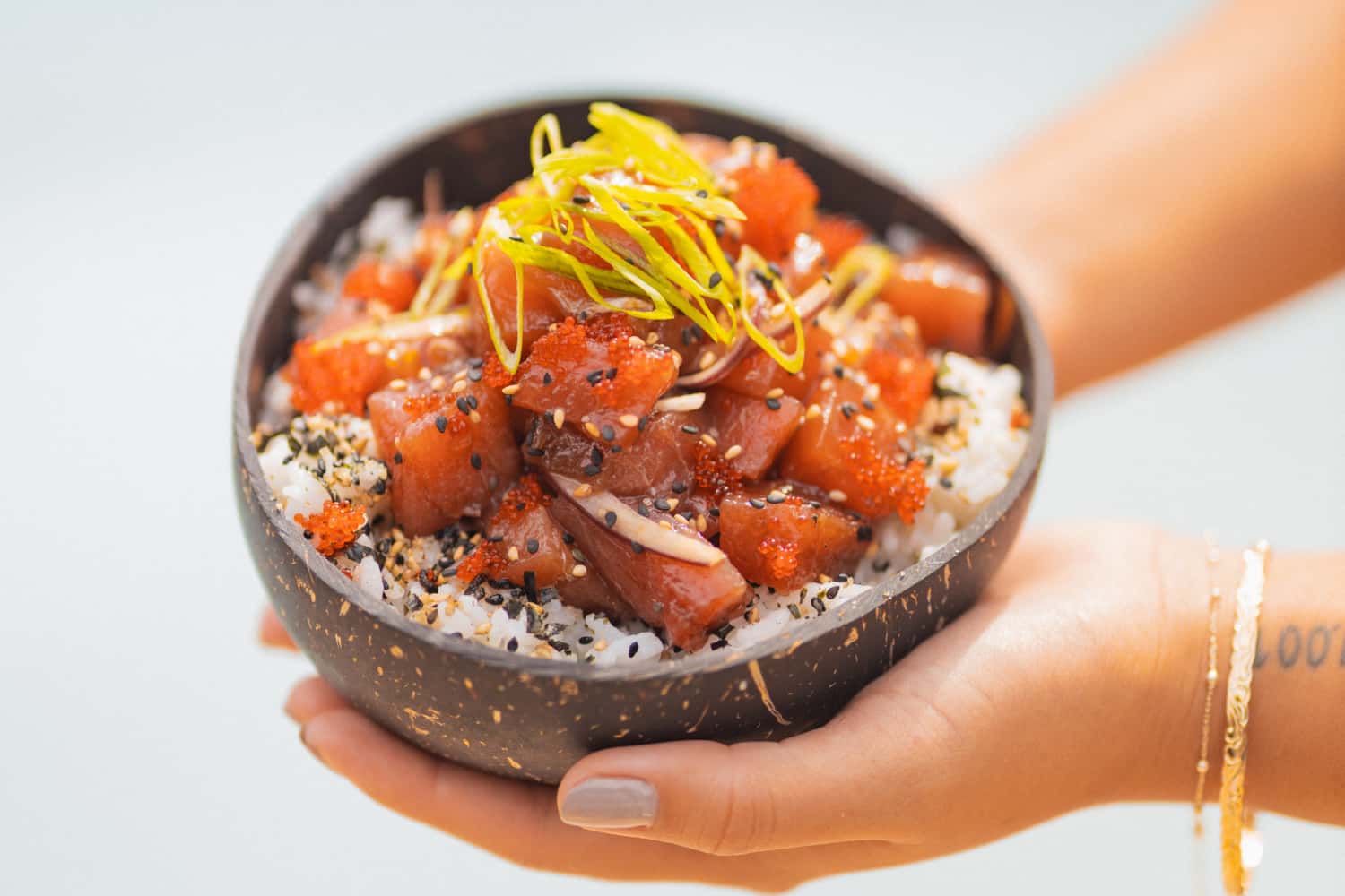 A bowl of poke and rice with furikake being held by two hands. The woman has on elegant gold bracelets.