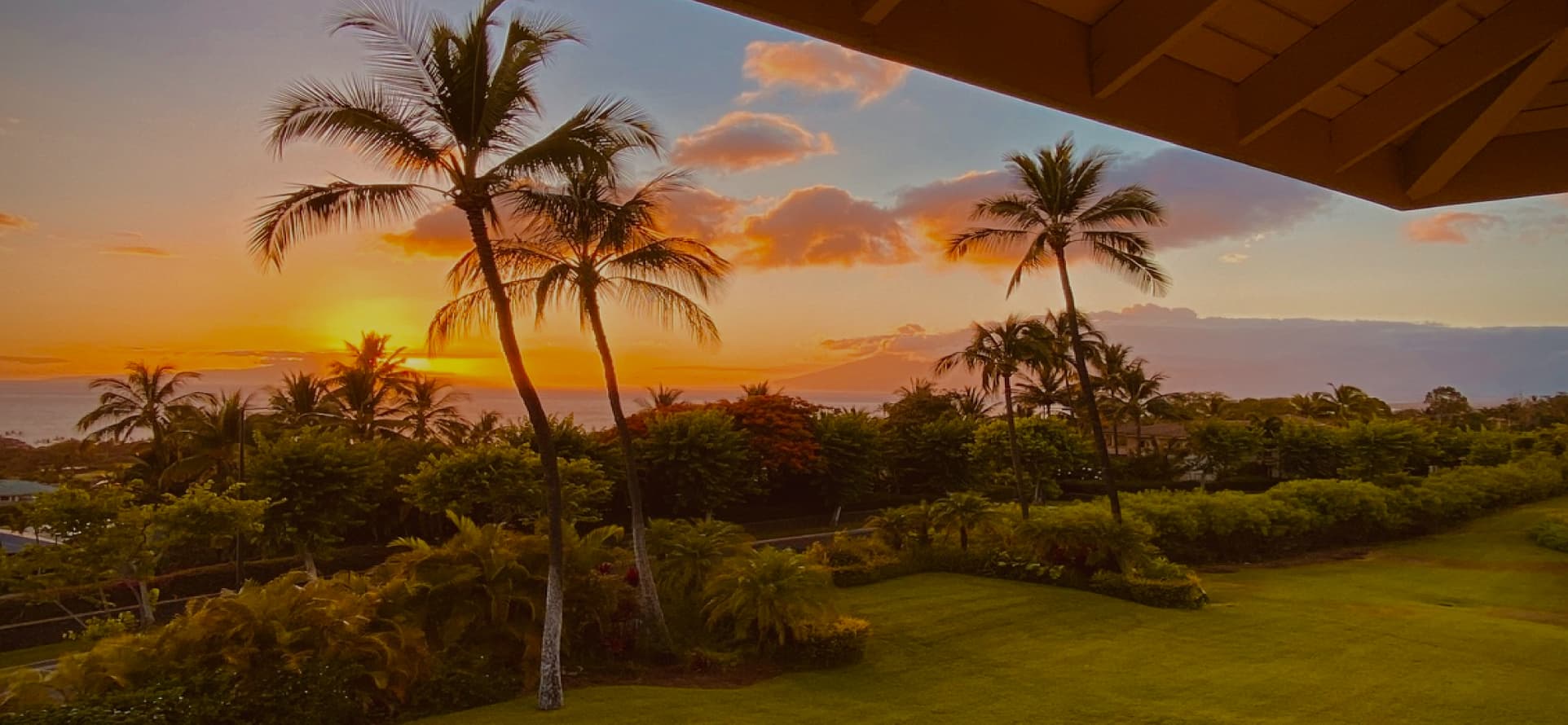 Sunset viewed from a veranda, with a golden sky above a silhouette of an island and the ocean. Two palm trees stand out against the vibrant colors. The foreground shows a neatly trimmed lawn and assorted tropical plants.