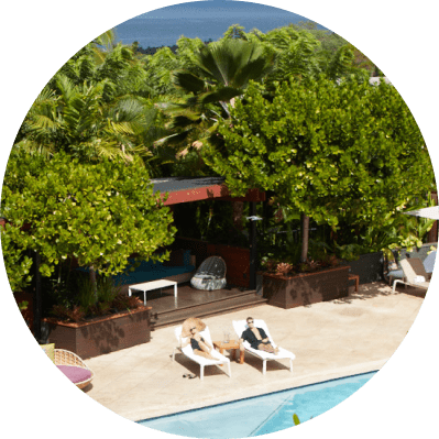 http://An%20aerial%20view%20of%20the%20pool%20surrounded%20by%20tropical%20trees.%20Two%20people%20are%20lounging%20on%20sunbeds,%20and%20there%20is%20a%20small%20building%20with%20a%20brown%20roof%20in%20the%20background.
