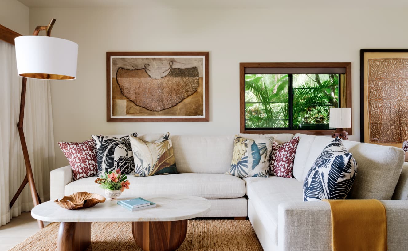 A modern living room featuring an off-white white couch with decorative pillows, a round coffee table with books on top, and a floor lamp. A framed abstract artwork hangs on the wall, and a window offers a view of greenery outside.