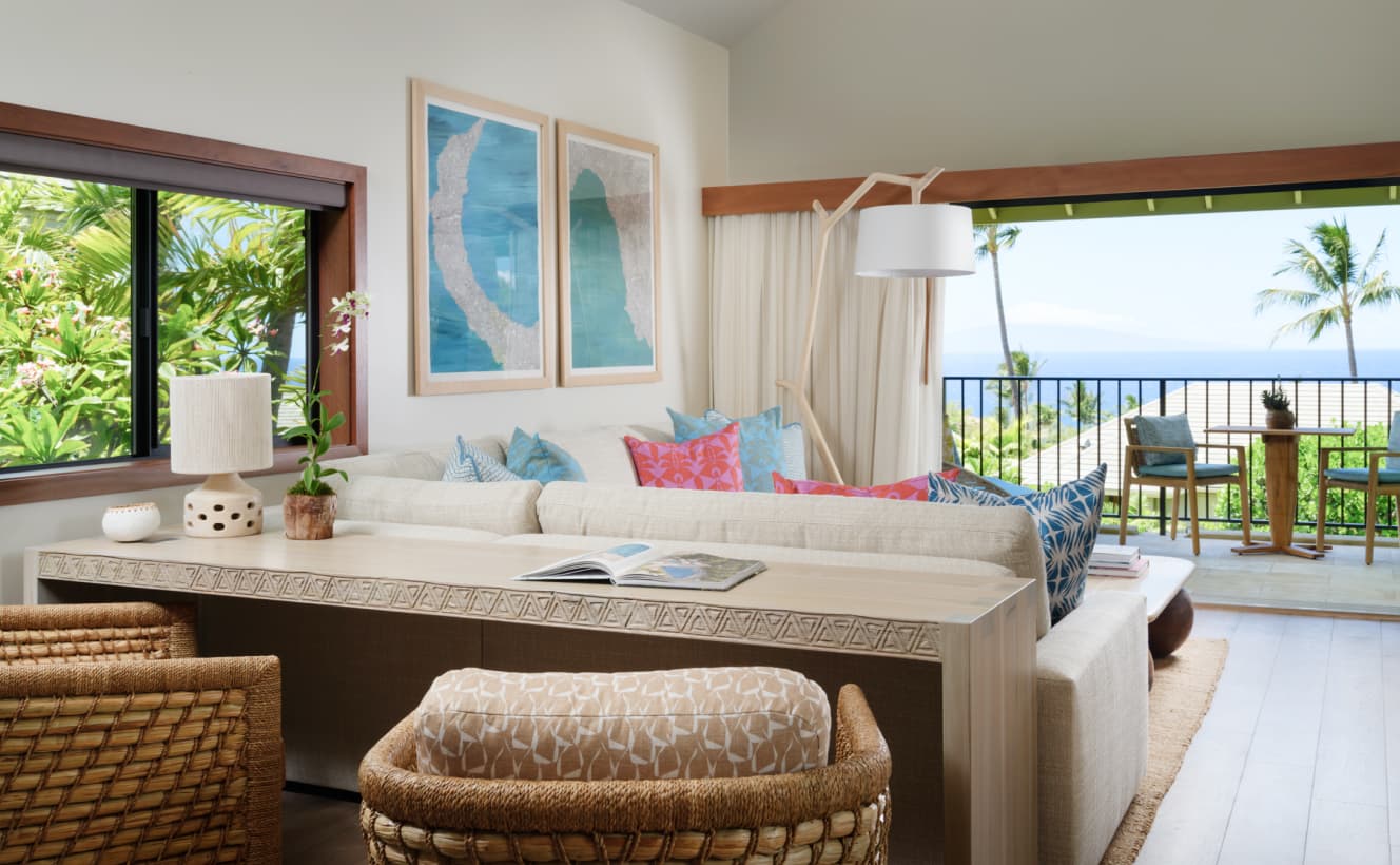 An open suite with a large off-white sofa filled with colorful cushions. In front of the sofa is a coffee table with a few books on top. The room has a warm, inviting feel with a large window that opens to a lanai overlooking the ocean.