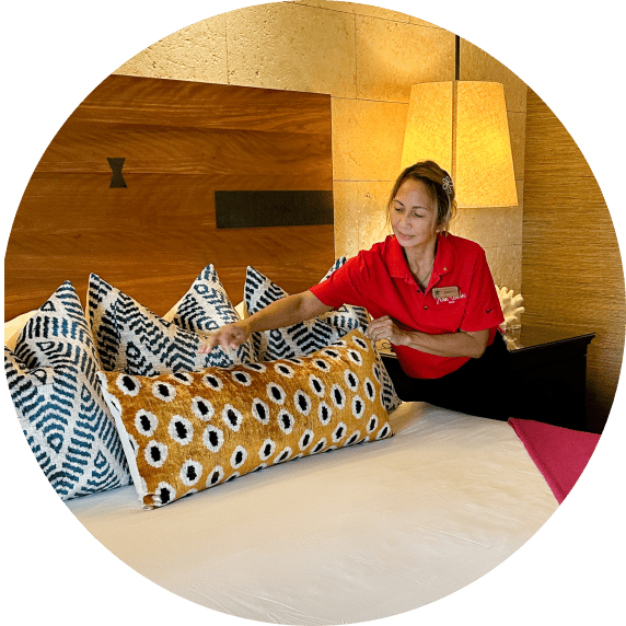 A hotel staff member in a red uniform is arranging decorative pillows on a bed. The pillows feature a mix of geometric and eye-catching patterns. Behind her is a stylish headboard and a lit table lamp, adding warmth to the room's ambiance.